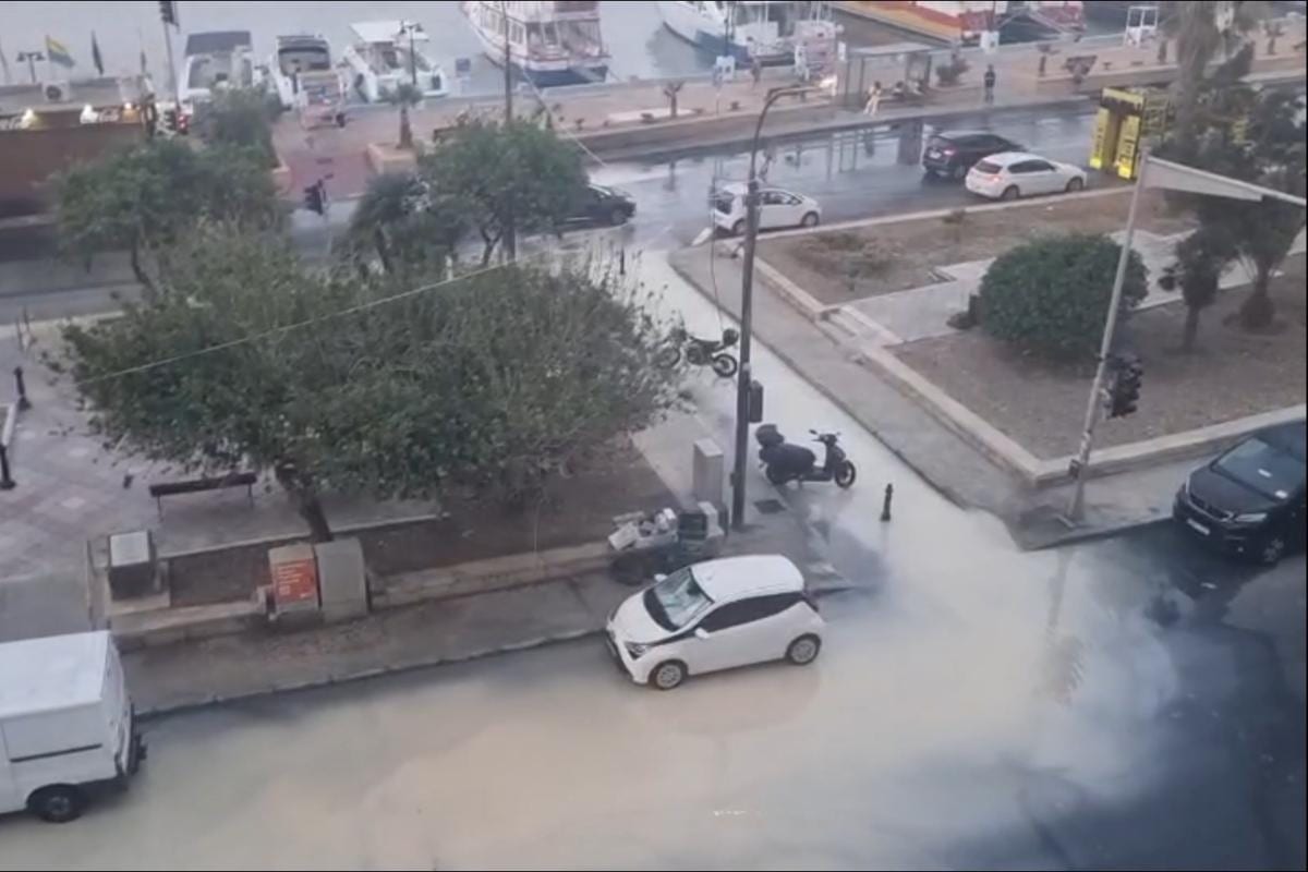 A flooded seafront square, brown water covering the road around parked cars and a scooter