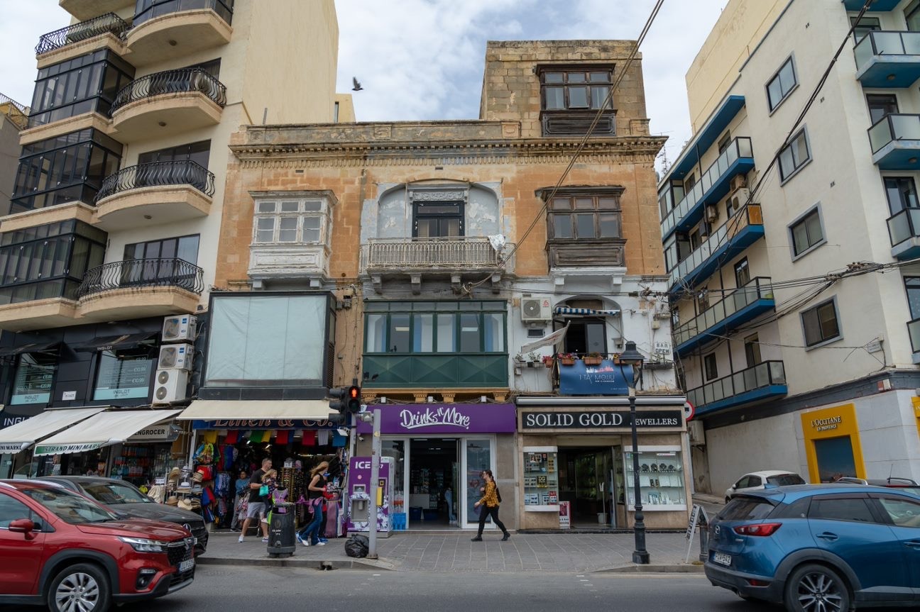 The 19th-century Sliema corner building before demolition: a traditional stone townhouse with an enclosed timber balcony and ground-floor shops, wedged between two taller modern apartment blocks on the seafront.