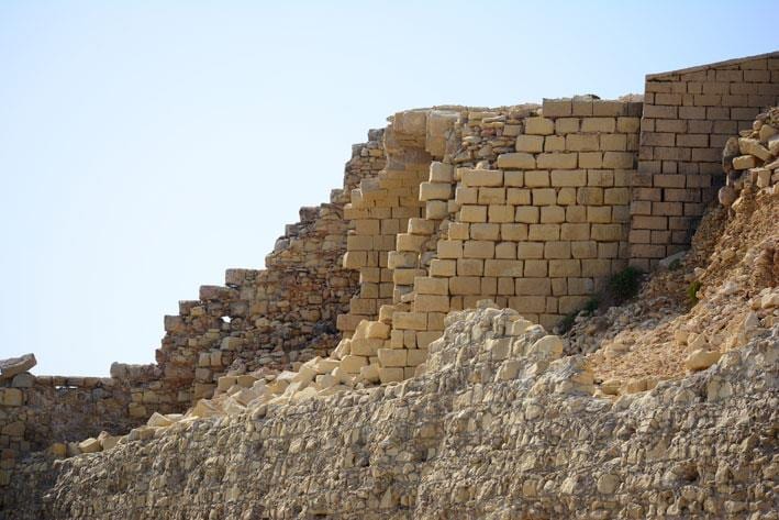 A collapsed section of Fort Ricasoli's fortification wall, dressed stone blocks tumbled against a clear blue sky.