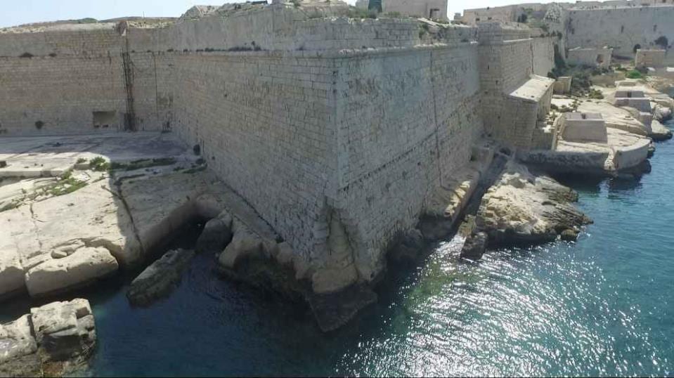 Aerial view of a massive limestone bastion of Fort Ricasoli rising straight out of the sea, its base undercut by the water, with the Grand Harbour fortifications behind.