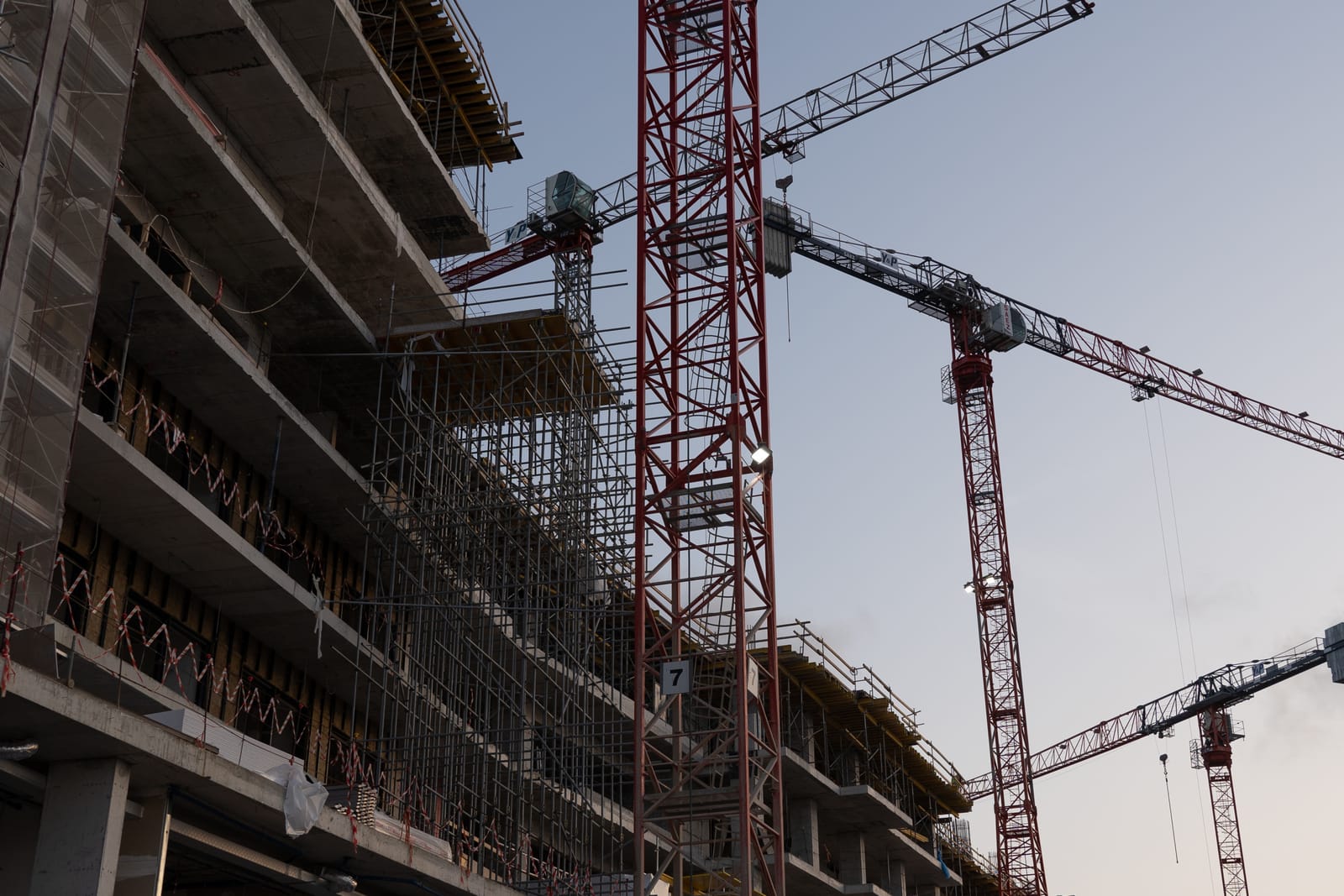 A raw concrete residential tower under construction, wrapped in scaffolding and surrounded by red cranes against a pale sky