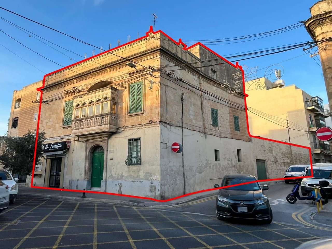 A two-storey limestone corner palazzo in Santa Venera with a traditional stone balcony and green shutters, the development site outlined in red, the aqueduct tower at the edge of the frame.
