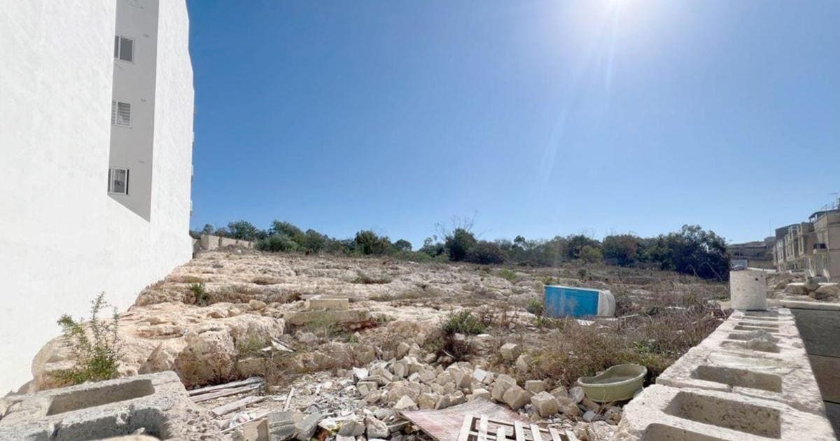 The rocky Kalkara site: rut-scored limestone strewn with rubble and a dumped bathtub, a blank apartment block wall rising at its edge